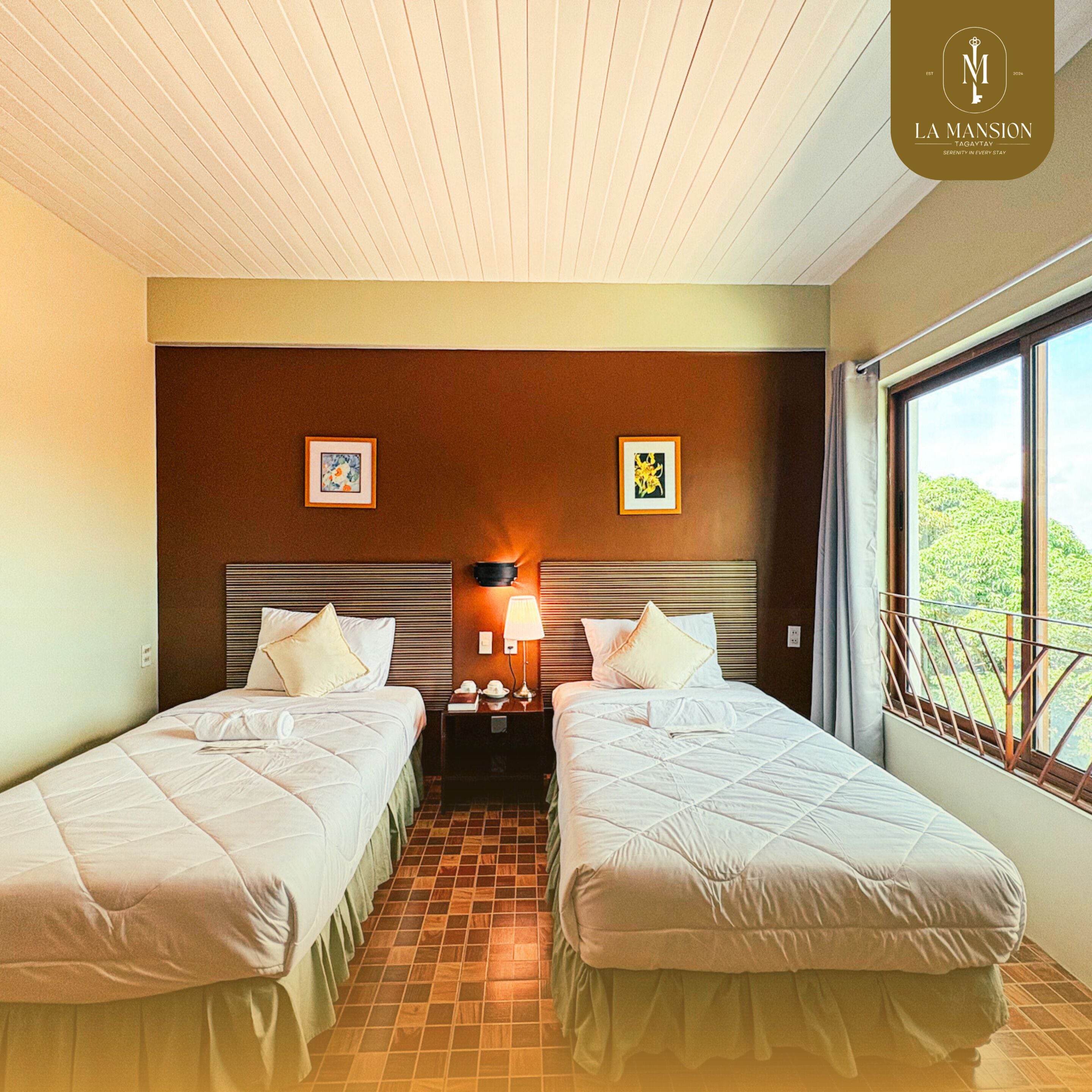 A hotel room with two beds, a nightstand with a lamp, framed artwork on the wall, and a window with a view of greenery.