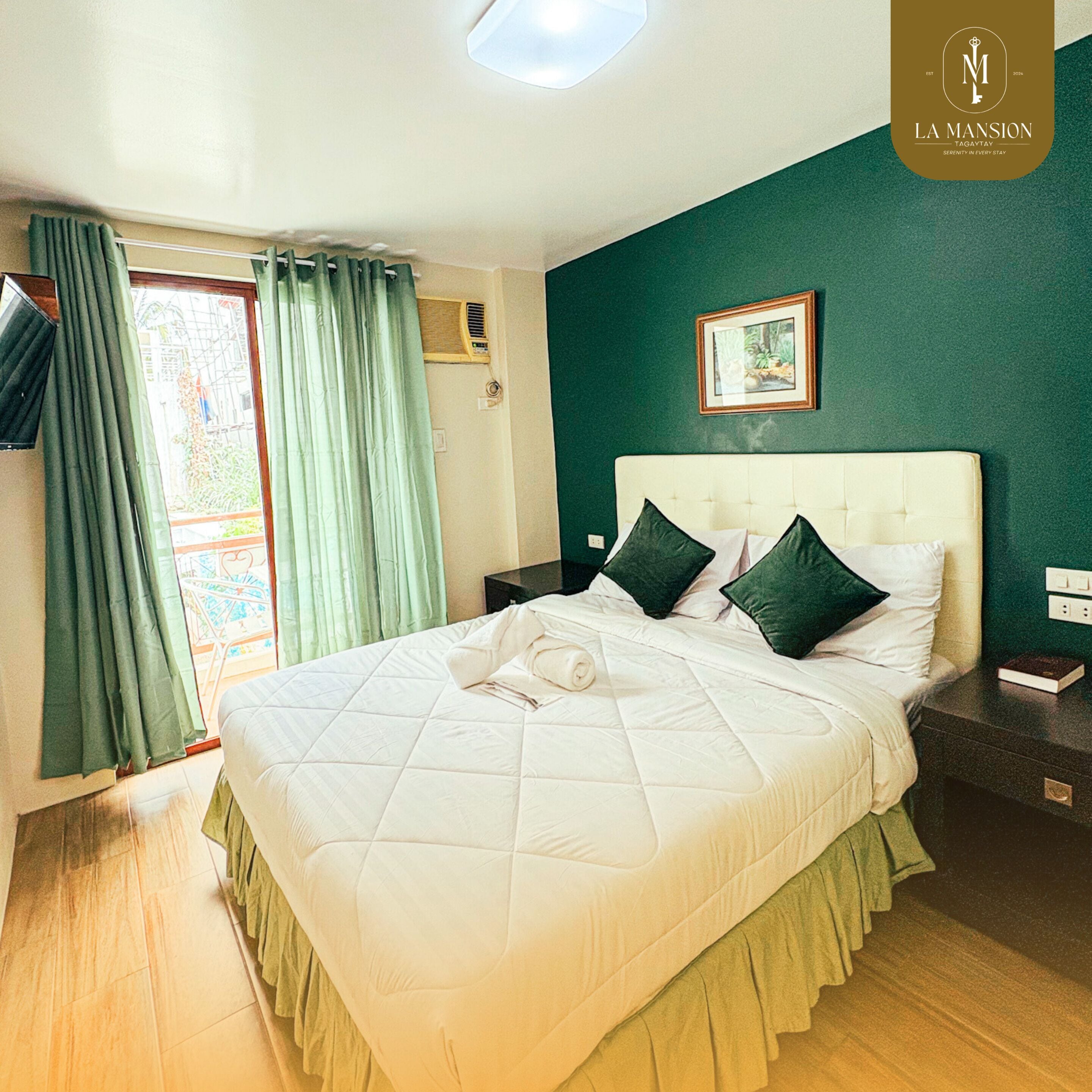 A neatly made bed with a quilted white duvet, two dark green pillows, and a folded towel. A framed picture hangs on the green wall above the bed.