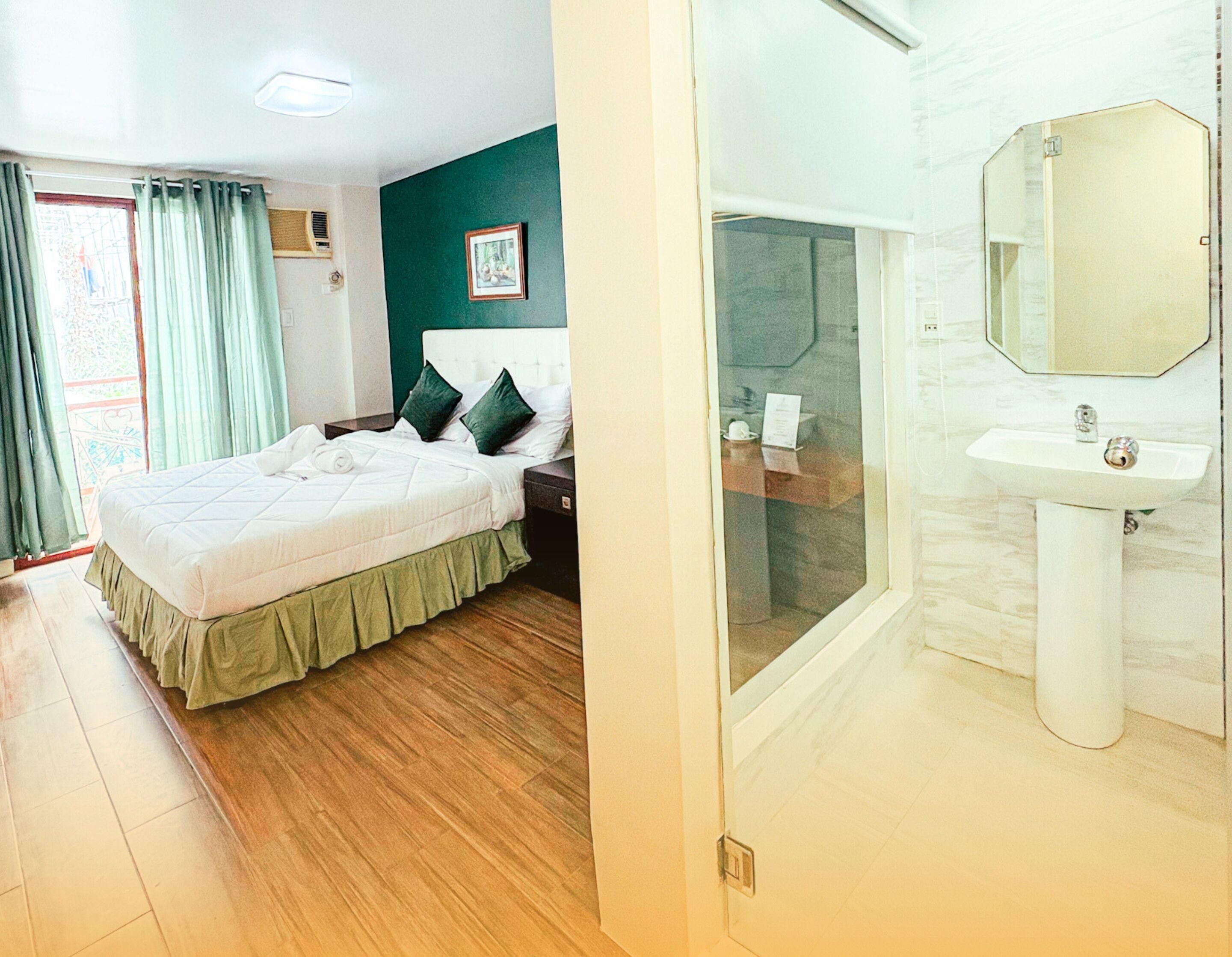 A bedroom with a bed, green curtains, and a nightstand. It also shows a bathroom with a sink, mirror, and shower.