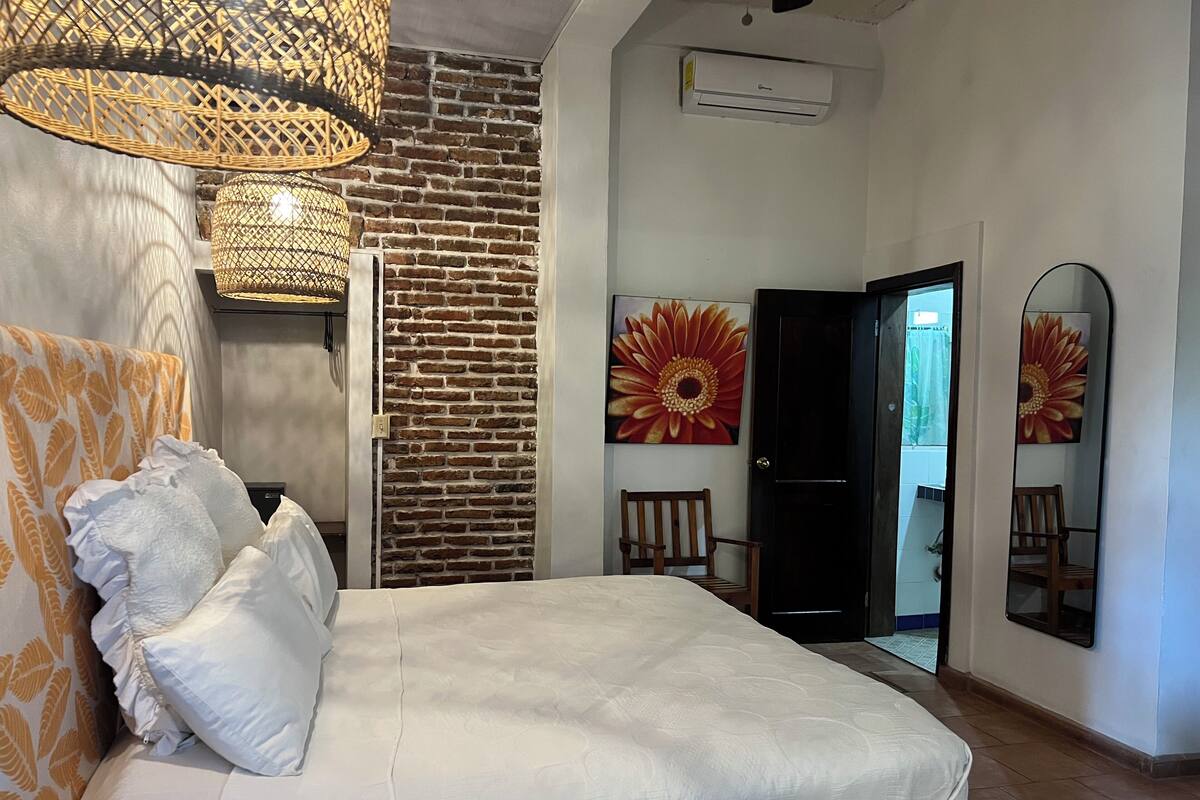 Quarto com parede de tijolos, cama com roupa de cama branca, duas luminárias de vime, um quadro grande de flores e uma porta que dá para outro cômodo.