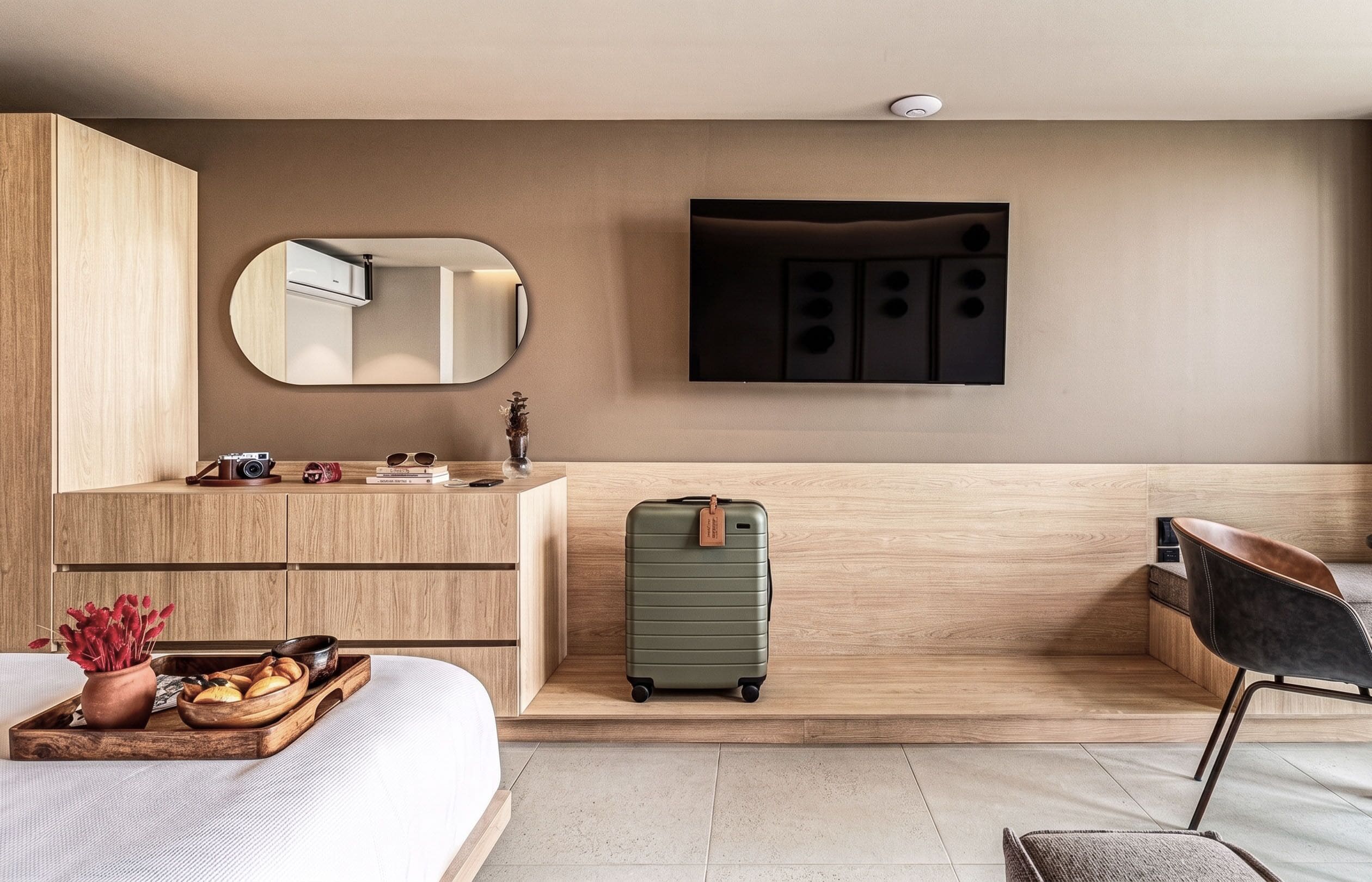Habitación de hotel moderna con tocador de madera, un espejo grande, televisor de pantalla plana, una maleta verde, cama y una silla.