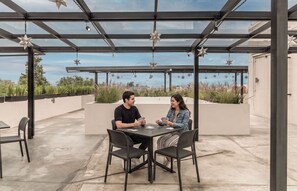 Un área de estar al aire libre con techo, una mesa y sillas, decoraciones colgantes con forma de estrella y vistas del cielo y edificios lejanos.