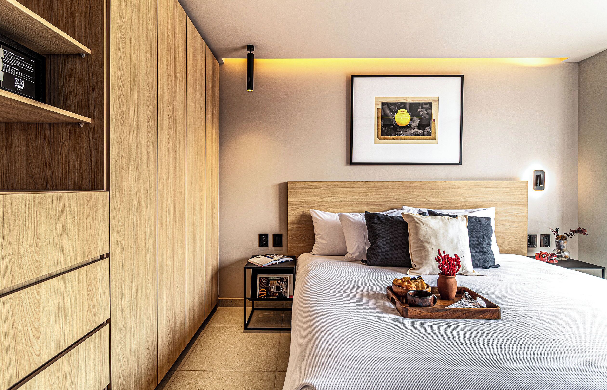 Une chambre d’hôtel avec une armoire en bois, un lit recouvert d’une courtepointe à rayures blanches et grises, un tableau encadré au mur et une petite table de chevet avec un livre et un vase de fleurs.
