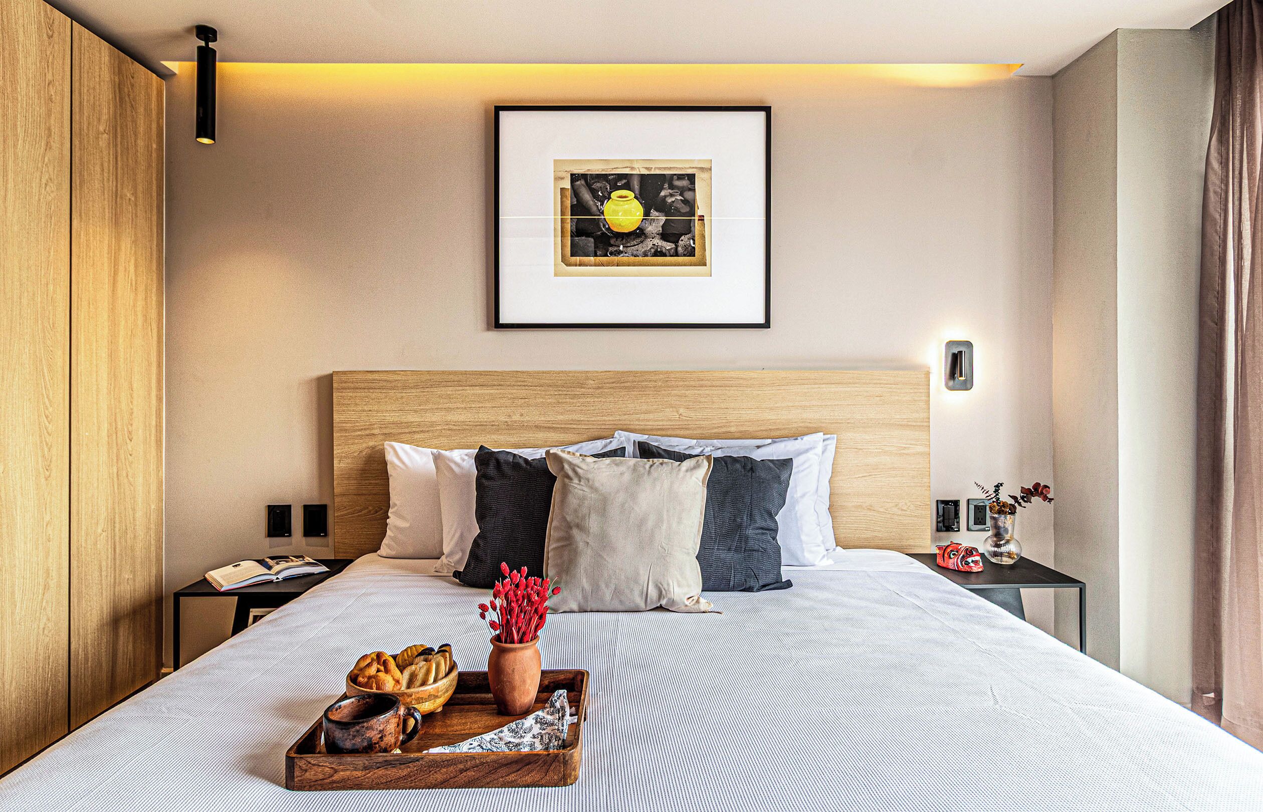 Habitación de hotel con una cama grande, un cuadro enmarcado en la pared, una mesita de noche con un teléfono y una bandeja con pan y un jarrón de flores.