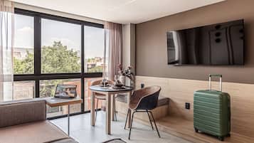 Une chambre d’hôtel moderne avec une grande fenêtre, une table à manger avec des chaises, une valise verte et un téléviseur à écran plat fixé au mur.