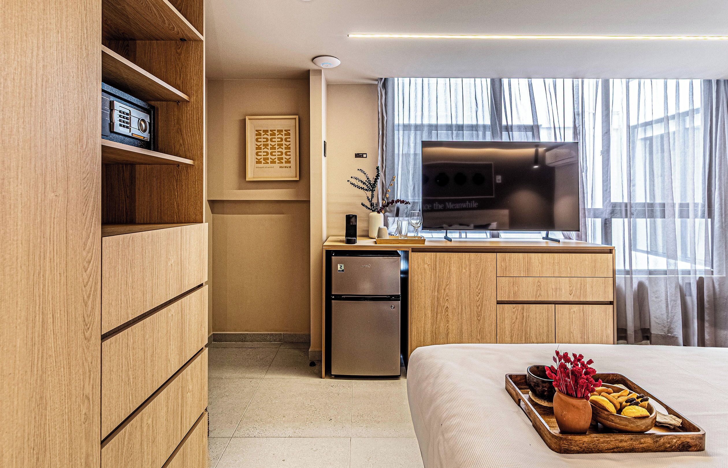 Quarto de hotel com cama, guarda-roupa de madeira, cozinha compacta com frigorífico, televisão e janela com cortinas.