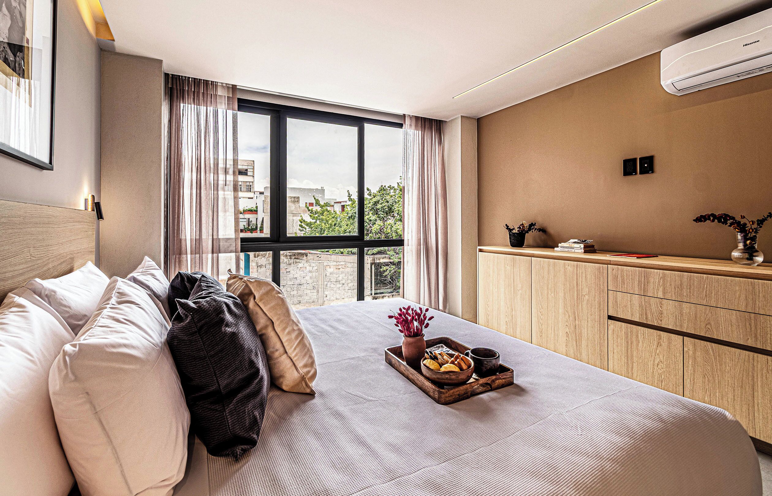 A hotel room with a bed, a bedside table with a tray of food, a vase with flowers, and a view of buildings through the window.