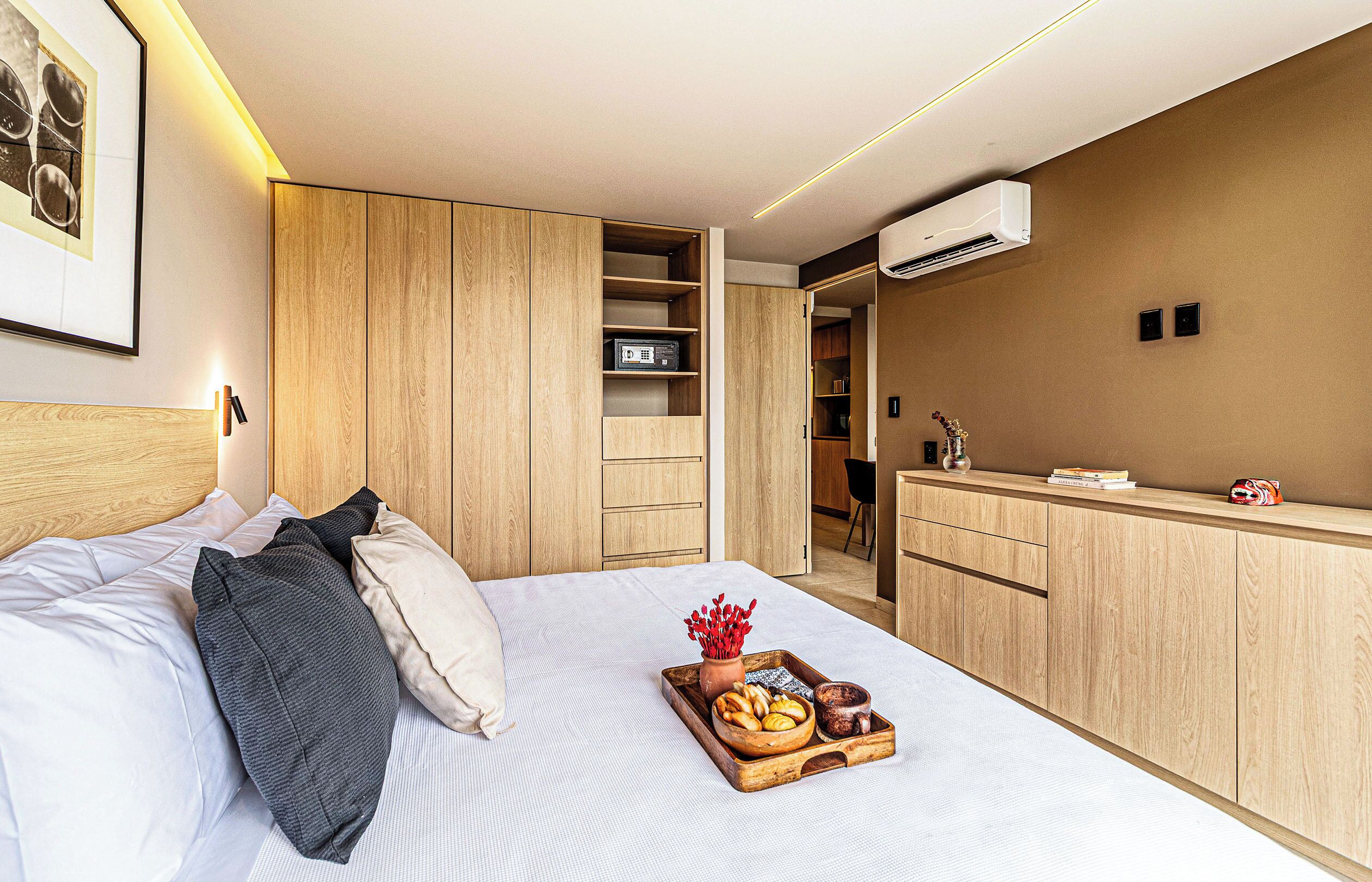 A hotel room with a bed, a bedside table with a tray of food, a wardrobe, a desk, and an air conditioning unit.