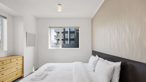 A modern hotel room with a large bed, a wooden dresser, a window with a view of buildings, and a textured wall. - First Tracks (Kutchan)