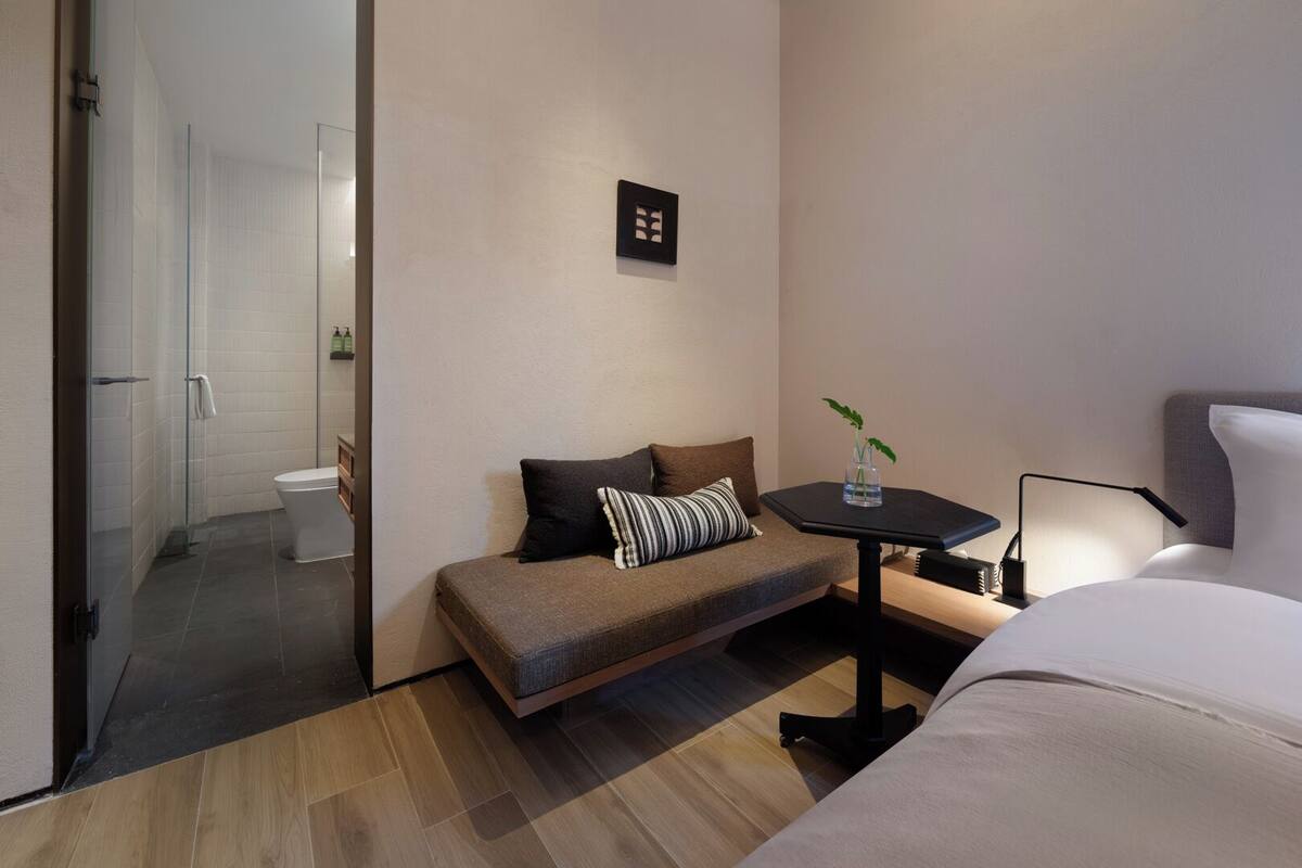 A modern hotel room with a sofa, a small table with a plant, a bedside lamp, and a bed.