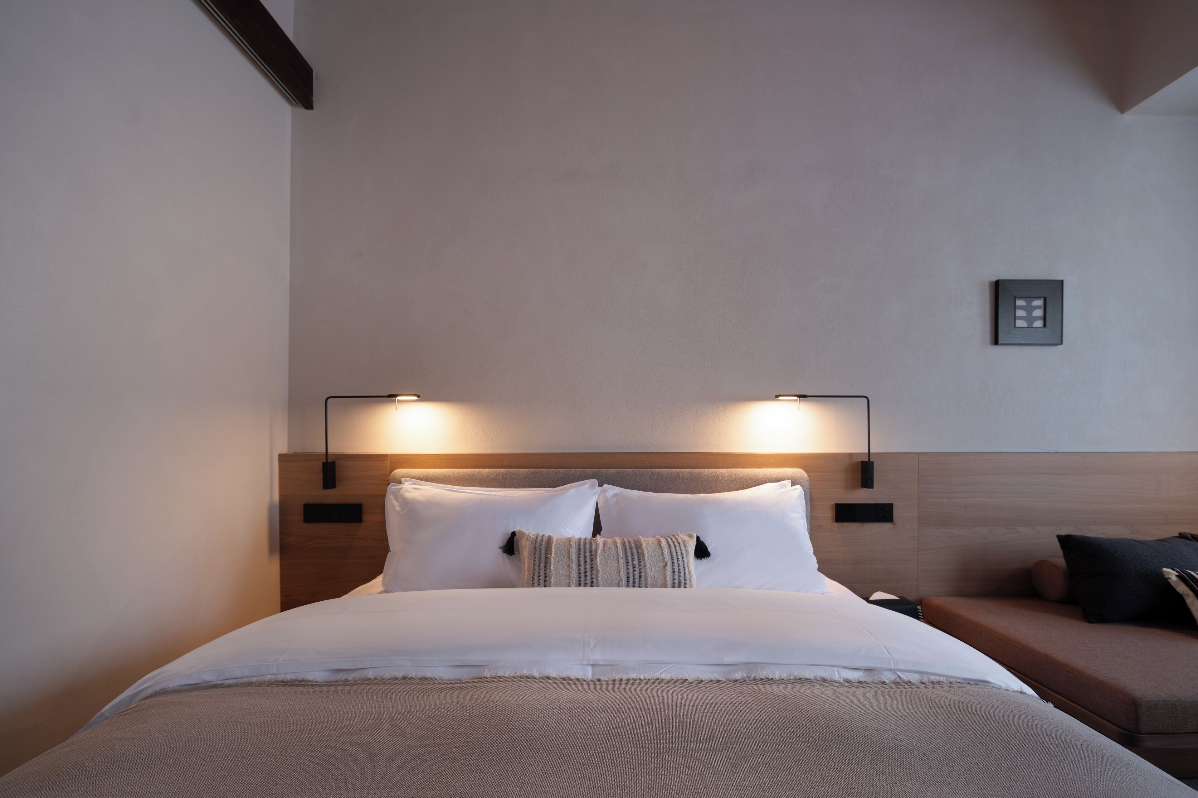 A neatly made bed with white linens and a striped pillow, a wooden headboard, and two wall-mounted reading lamps.