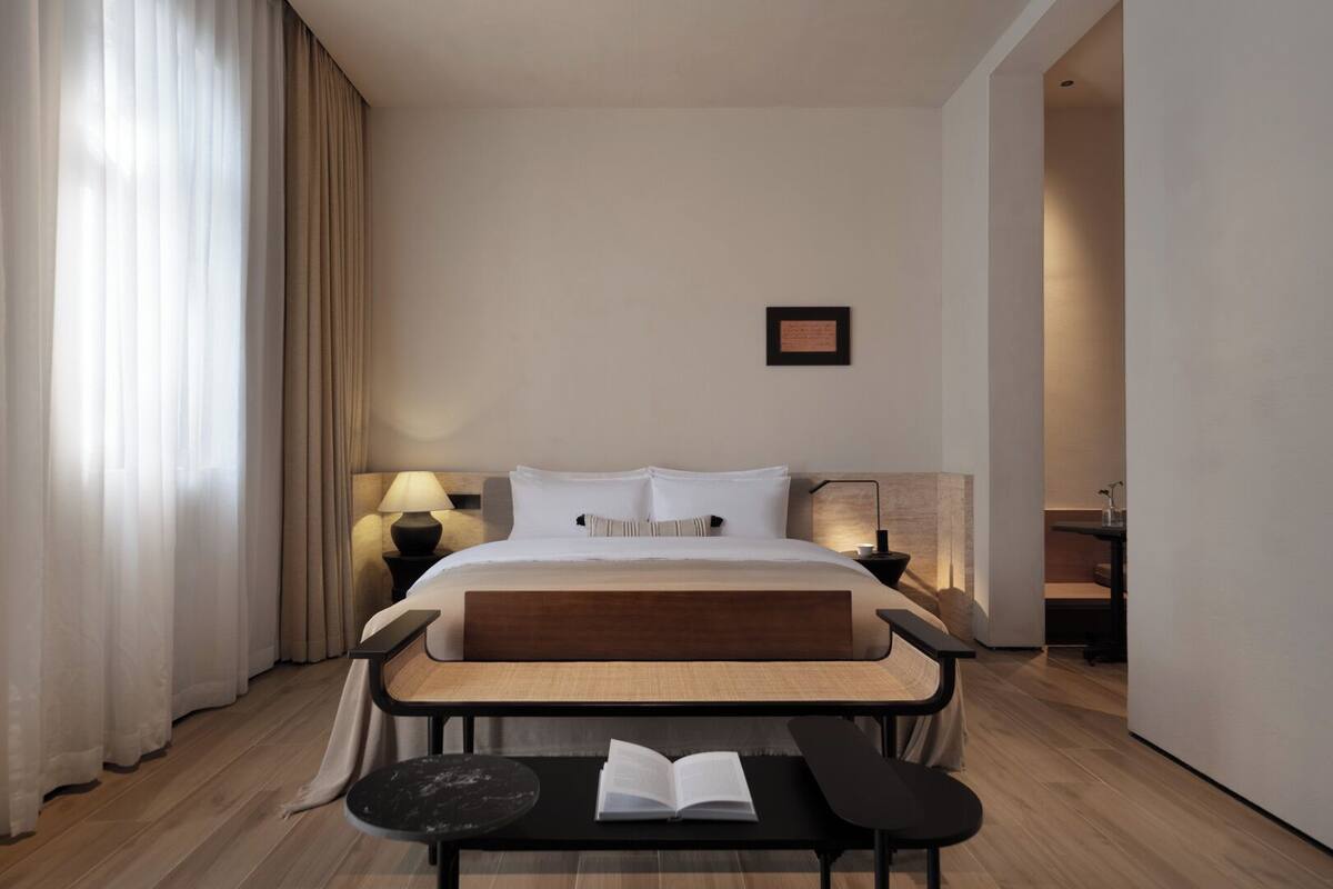 A modern hotel room with a large bed, bedside lamps, a small table with a book, and a bench.