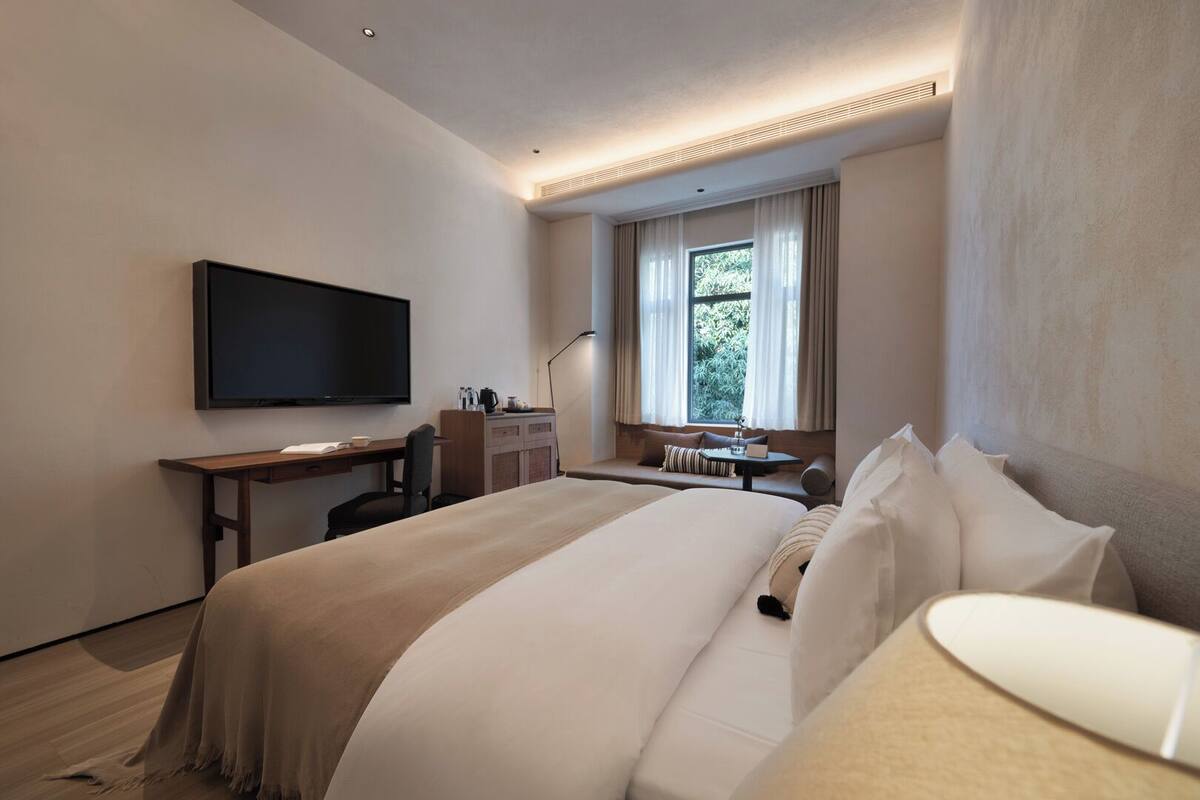 A hotel room with a large bed, a desk with a chair, a flat-screen TV, a window with curtains, and a view of greenery outside.