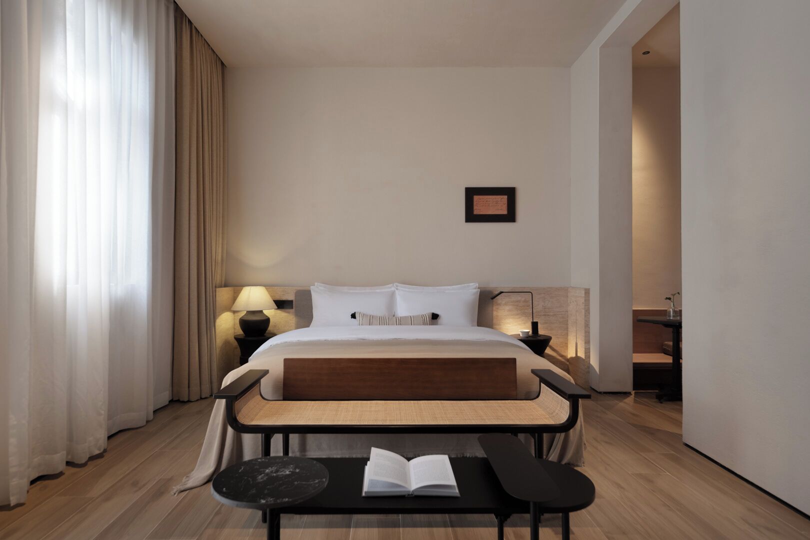 A modern hotel room with a large bed, bedside lamps, a small table with an open book, and a bench.