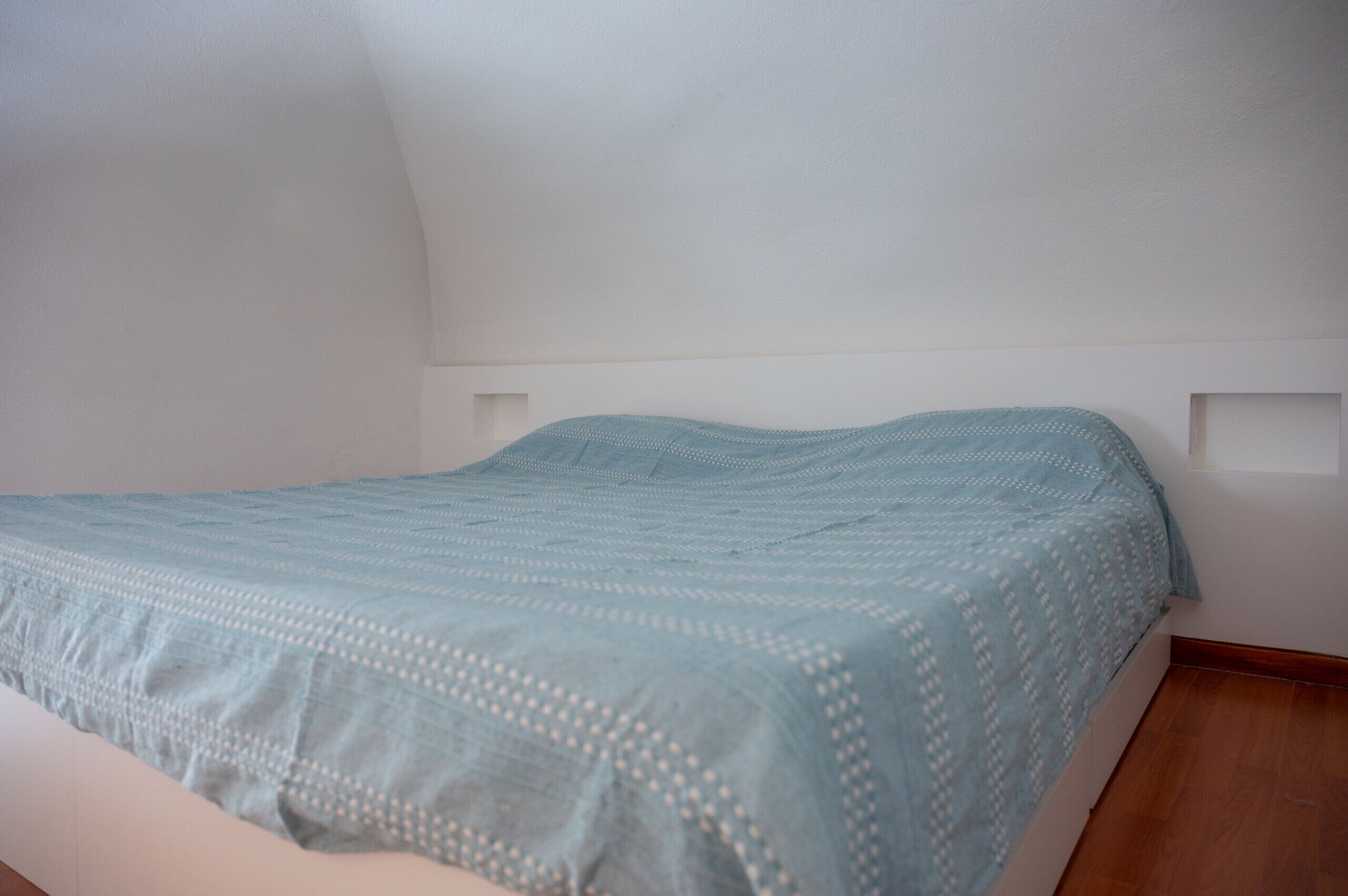 A simple, unadorned bedroom with a single bed and a plain white wall.