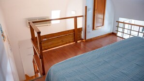 Une chambre avec un escalier en bois, un lit recouvert d’une couverture bleue et une fenêtre avec un cadre en bois.