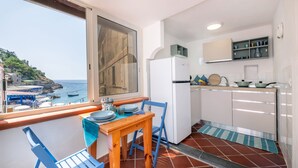 Un balcon avec une table et des chaises en bois, une vue sur la mer, et une cuisine avec des meubles blancs et un réfrigérateur.