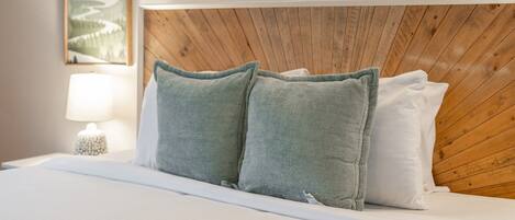 A neatly made bed with green and white pillows, a wooden headboard, a bedside lamp, and a framed picture on the wall.