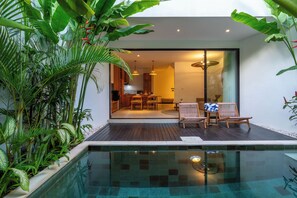 Un espace piscine moderne avec une terrasse en bois, des chaises longues et un coin repas fermé par des parois de verre.
