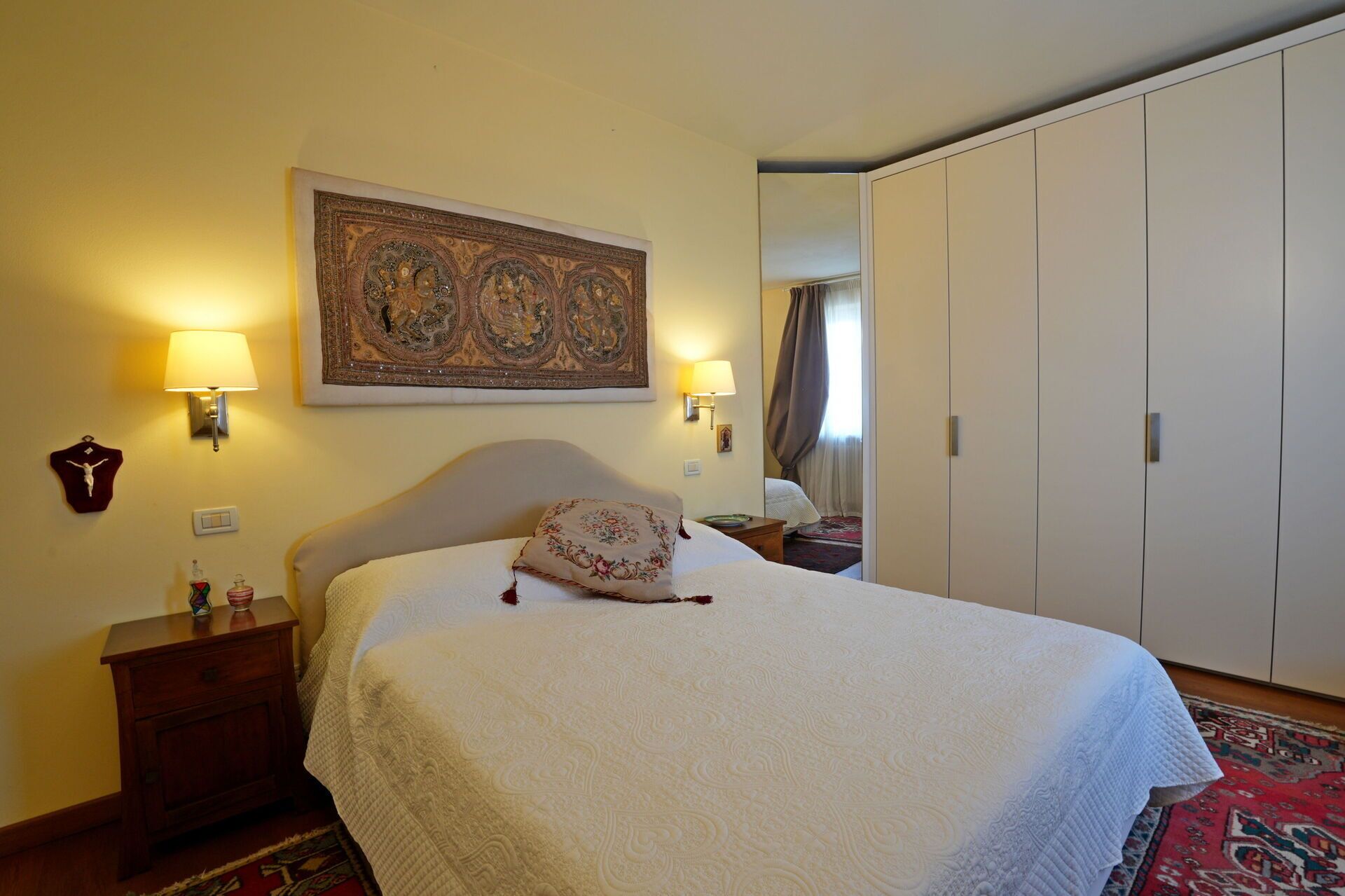 Une chambre d’hôtel avec un grand lit, deux lampes de chevet, une tapisserie décorative, une armoire et une table de chevet avec une bouteille et un verre.