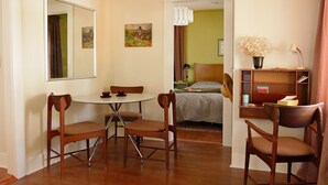 A hotel room with a dining table, two chairs, a bed, a nightstand, a lamp, a bookshelf, and a mirror.