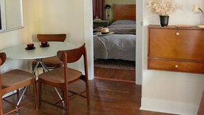 Una habitación con una mesa de comedor y sillas, una cama con cabecera, una mesita de noche con lámpara, un aparador y un cuadro en la pared.