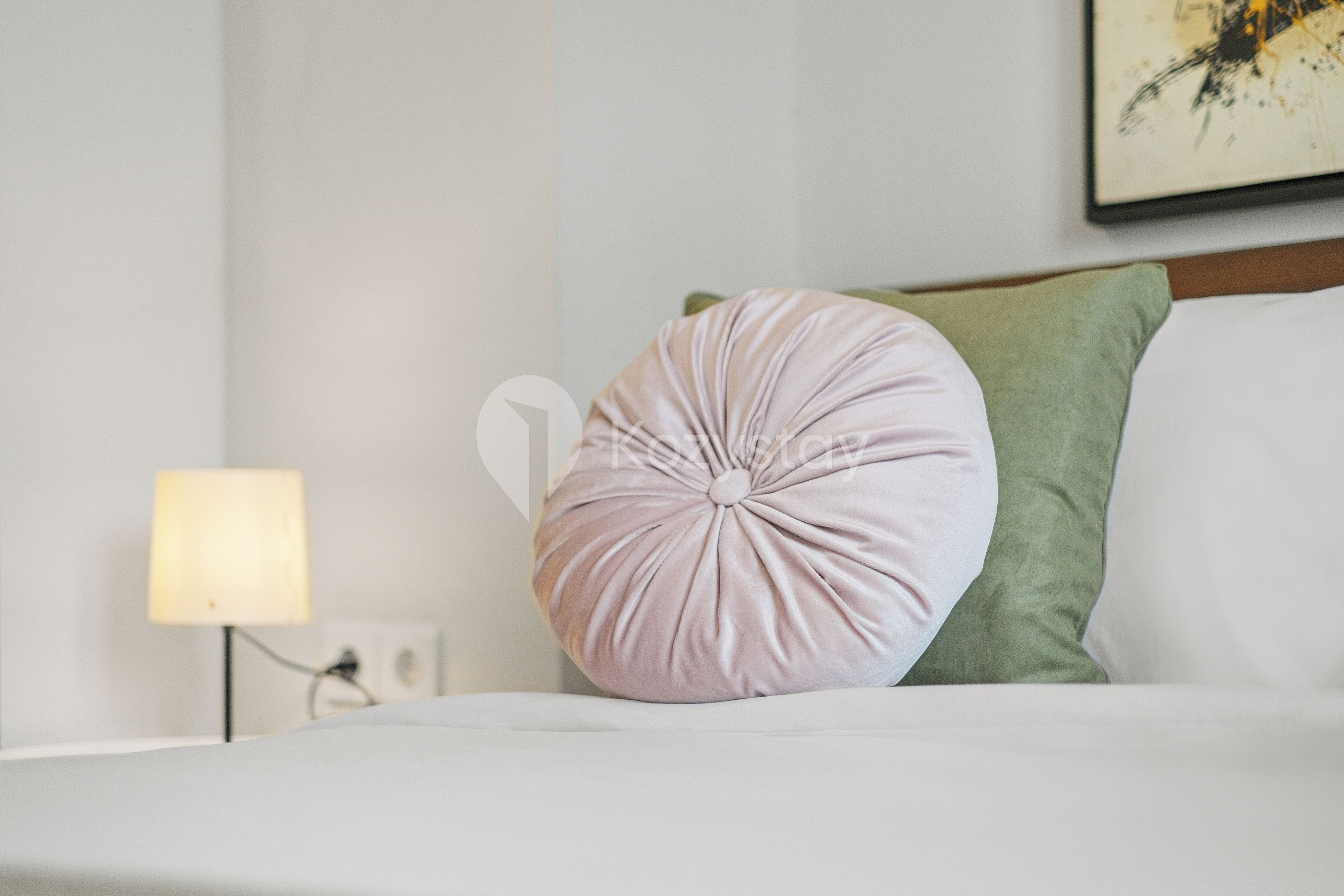 Una cama bien tendida con un almohadón redondo de color claro y una almohada verde. Una lámpara está sobre una mesita de noche a la izquierda.