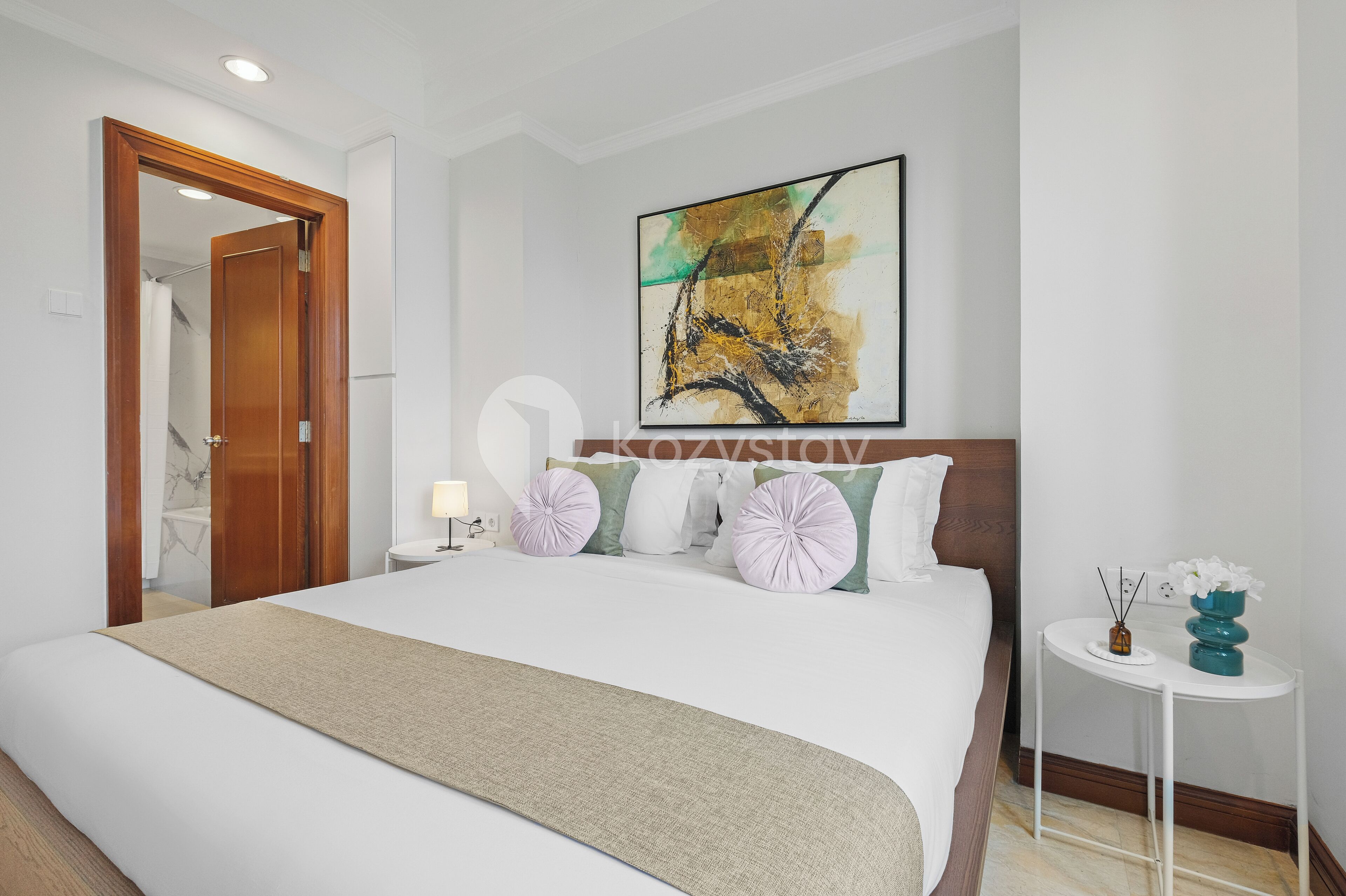 A hotel room with a large bed, a wooden door, a painting on the wall, and a small table with a vase.