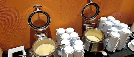 En buffet med to store gryder mad, flere hvide kopper og en lille skål med sauce.
