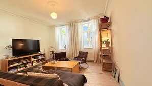 Ett vardagsrum med en tv, en soffa, ett soffbord och bokhyllor. Det finns två fönster med gardiner, ett trägolv och en liten hylla med en växt.