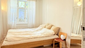 Ett sovrum med en säng i trä, en stol, ett fönster med gardiner och en spegel.