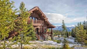 A log cabin with a balcony, surrounded by snow-covered ground and pine trees, with mountains in the background. - Vast Ski In/Ski Out Big Sky Chalet (Big Sky)