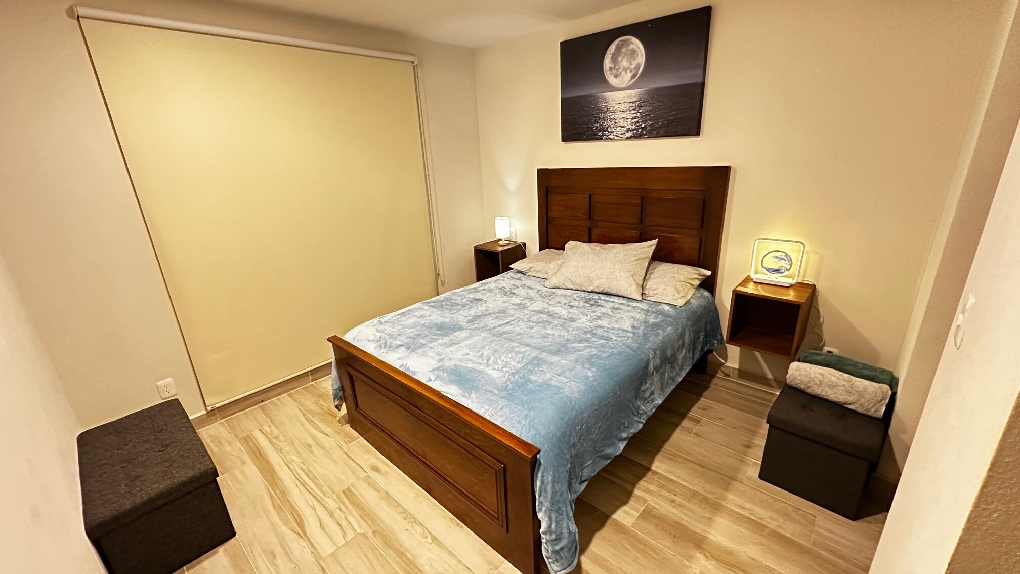 Un dormitorio con cama de madera, una mesita de noche con lámpara, una pequeña mesa auxiliar con lámpara, un cuadro en la pared y un armario.