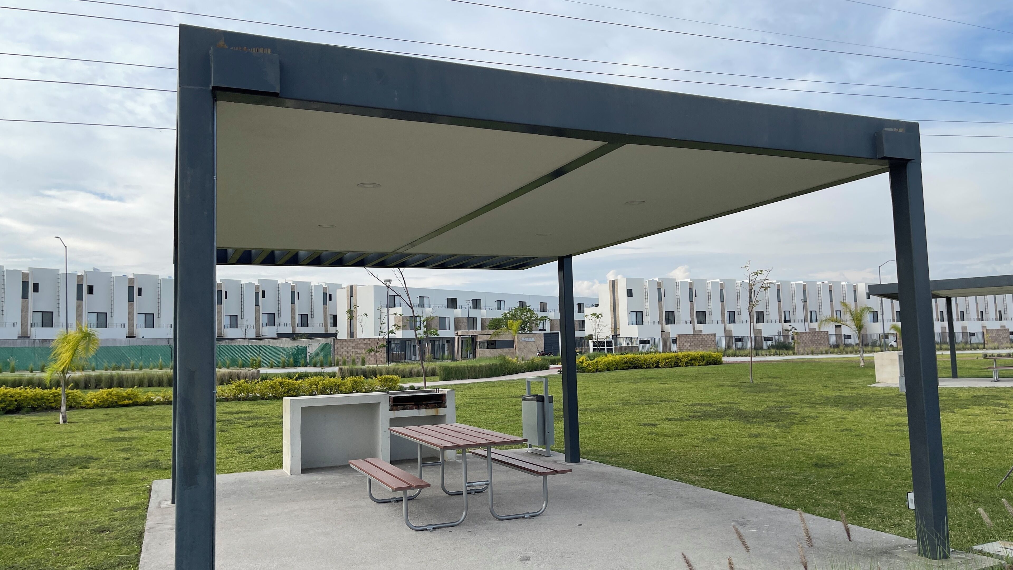 Un espacio al aire libre cubierto con una mesa de picnic y bancos, rodeado de un césped cuidado y una hilera de edificios modernos al fondo.