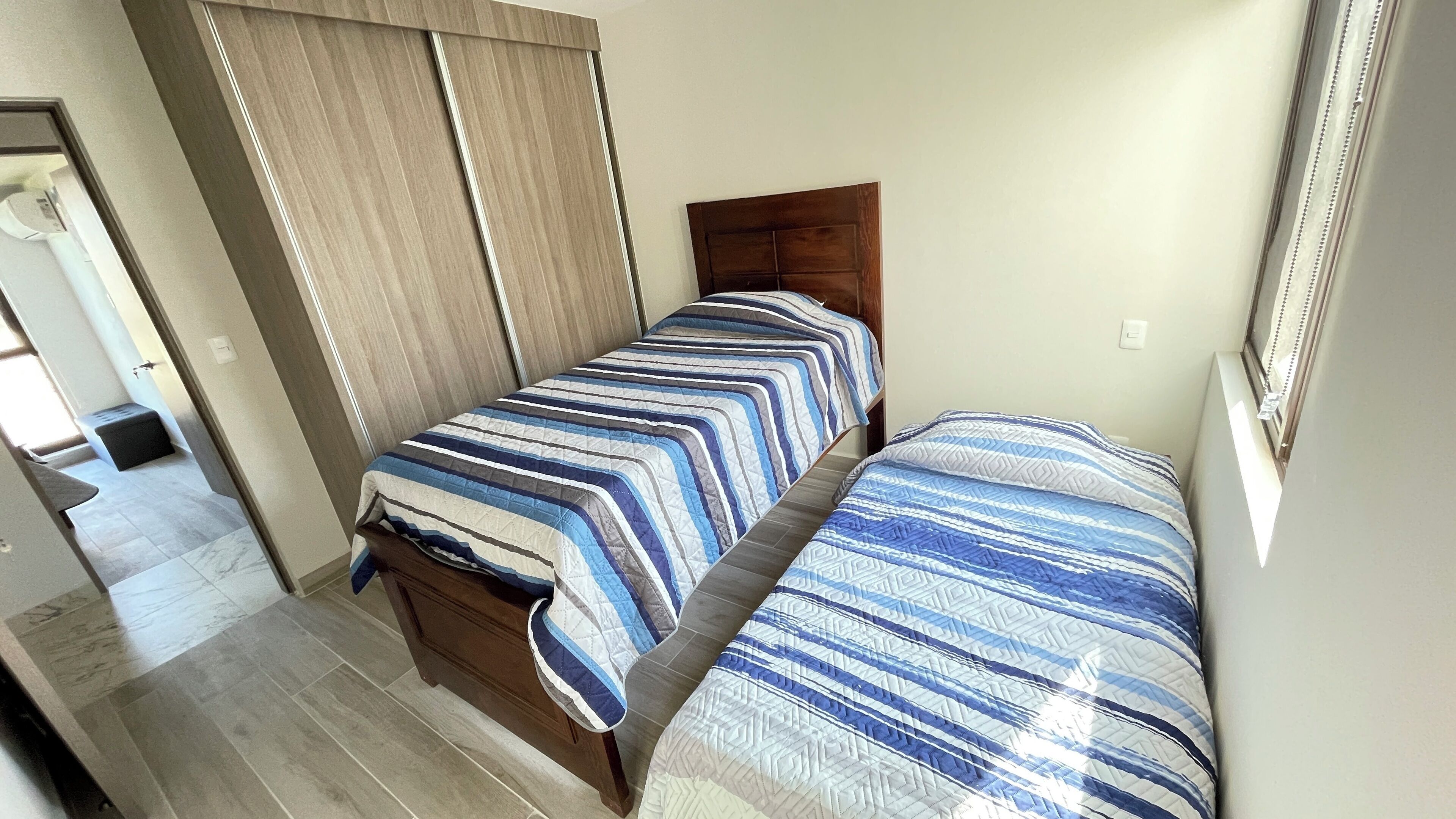 A small, neatly arranged bedroom with two single beds, a wooden headboard, and a tiled floor.
