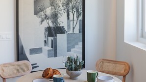 A dining area with a round table, two chairs, a framed black and white artwork, a cactus plant, a croissant, and two cups.