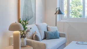 Una sala moderna con un sofá beige, una mesa auxiliar redonda de madera, una lámpara de pie y un ventanal con vistas a árboles.