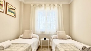 Una habitación con dos camas, una mesita y un ventanal con cortinas traslúcidas.