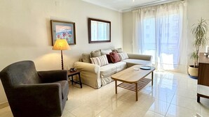 Una sala de estar con sofá, sillón, mesa de centro y lámpara. Se observan dos cuadros en la pared, una ventana con cortinas y una planta en maceta.