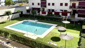 Una piscina rodeada de un césped cuidado, con un edificio al fondo que tiene balcones y toldos rojos.
