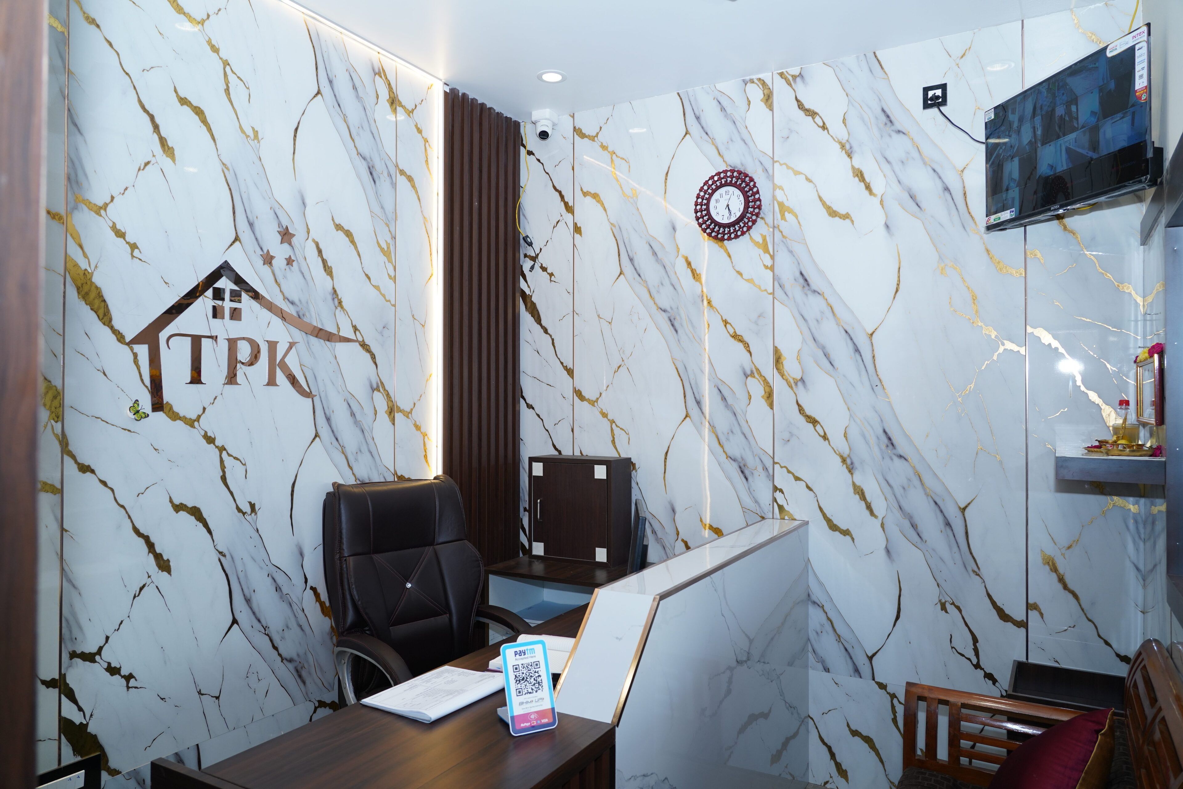 Una oficina moderna con pared de mármol, el logotipo de TPK, un escritorio con silla, un televisor y un reloj.