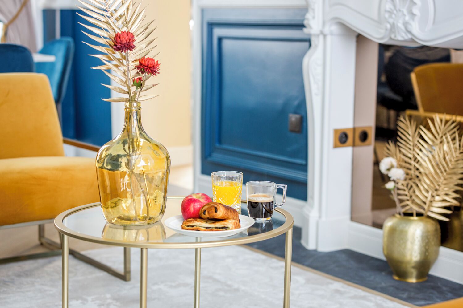 Ein Esstisch mit einer Flasche, einem Teller mit Gebäck, einem Glas Orangensaft und einer Kaffeetasse in einem Zimmer mit blauen und gelben Elementen.