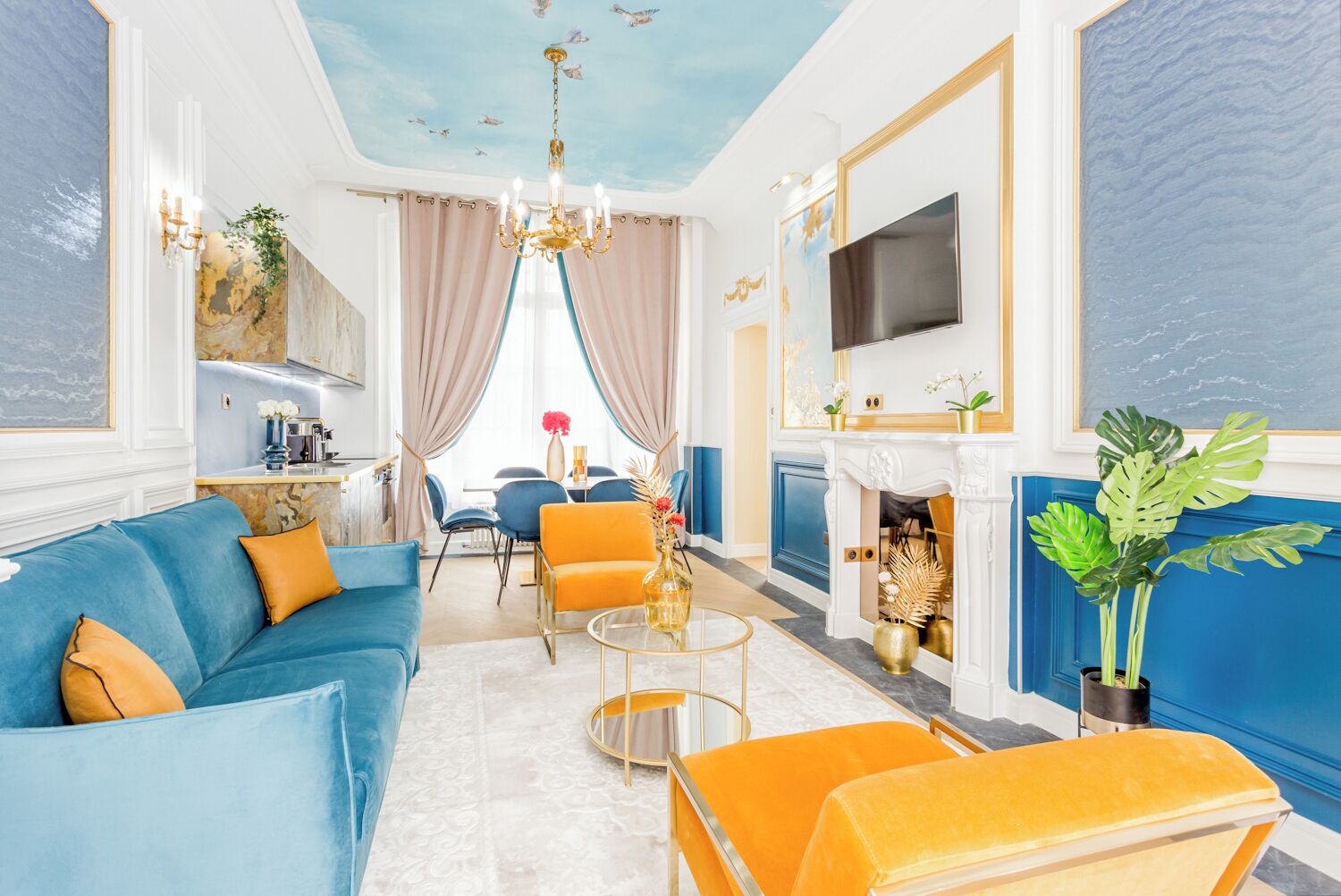A spacious living room with a blue sofa, orange armchairs, a fireplace, and a TV mounted on the wall.
