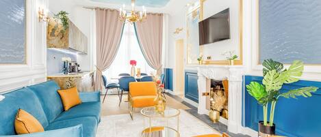 A spacious living room with a blue sofa, orange armchairs, a fireplace, and a TV mounted on the wall.