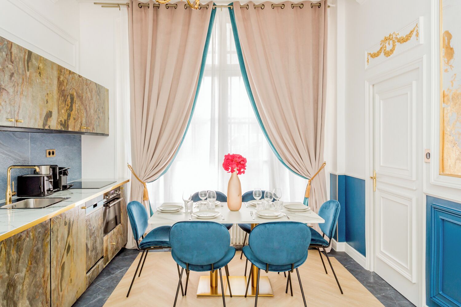 Un coin repas avec une table blanche dressée pour quatre personnes, des chaises bleues, un vase avec des fleurs rouges et une cuisine dotée d’un dosseret en marbre et de robinetterie dorée.
