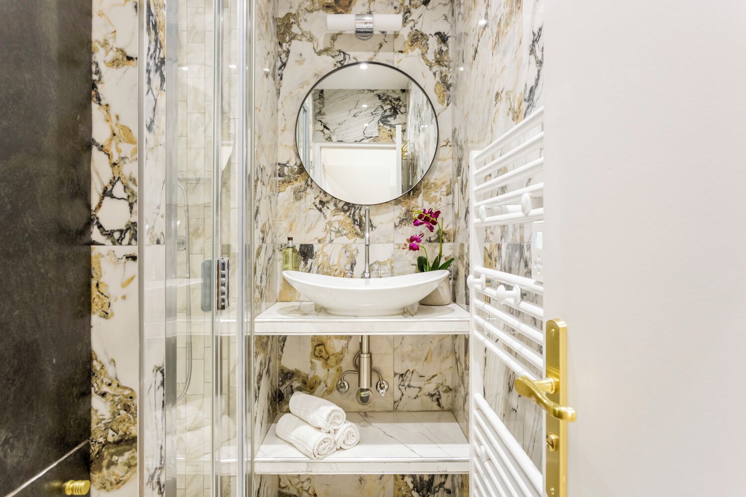 A bathroom with a glass-enclosed shower, a white sink with a marble backsplash, and a gold door handle.