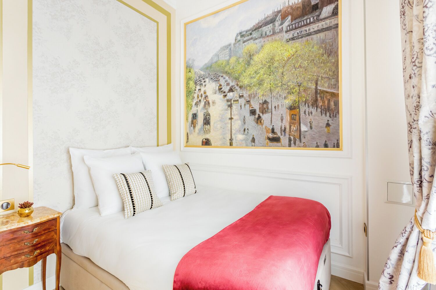 A hotel room with a bed, a nightstand, a lamp, and a painting of a street scene.