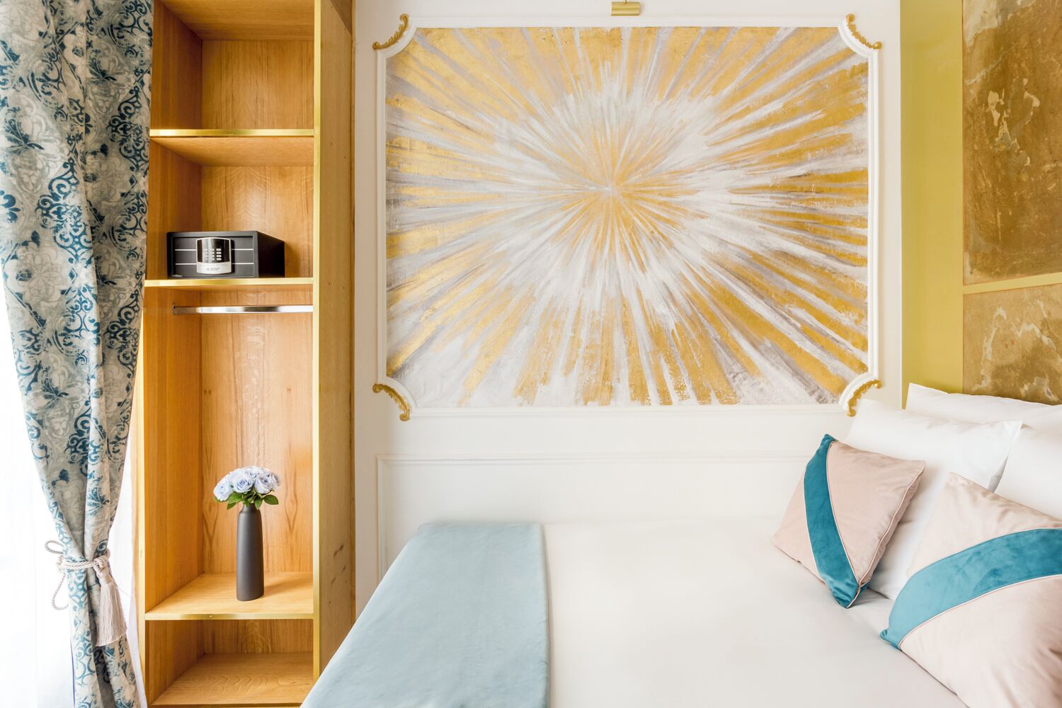 Une chambre à coucher avec un lit, une étagère sur laquelle se trouve une radio, un vase avec des fleurs et un grand tableau abstrait accroché au mur.