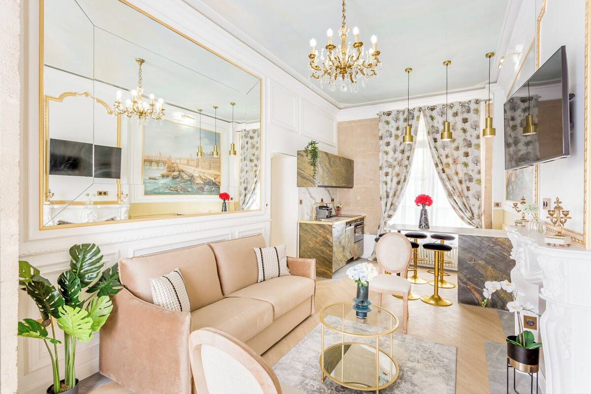 A spacious living room with a beige sofa, a coffee table, and a dining area with a marble island. A large mirror and chandelier are visible, along with a television and a kitchen area in the background.