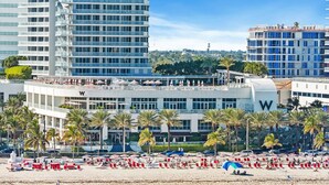 Un hotel frente a la playa con varios pisos, un llamativo letrero con la letra 'W' y una zona de playa con tumbonas y sombrillas.